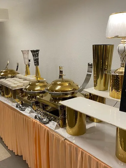 A close-up of our main course station at an indoor event. A clear sign indicates the course, and the arrangement of gleaming gold chafing dishes and decorative vases showcases our organized and elegant approach.