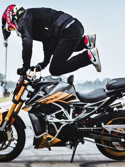 A rider performs a "Christ" stunt, standing on the tank of their bike with arms outstretched. This is a classic, high-skill move that always wows the crowd.