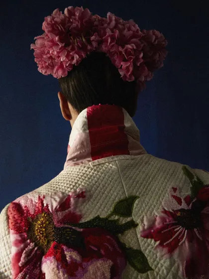 A detailed shot of a reversible quilted jacket with intricate floral embroidery. The styling focuses on the craftsmanship of the garment, with a floral headpiece creating a cohesive, art-inspired look against a solid blue background.