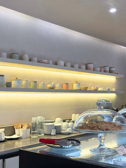A look at our counter, with cookies under a glass cloche and our collection of ceramic cups neatly arranged on backlit shelves.
