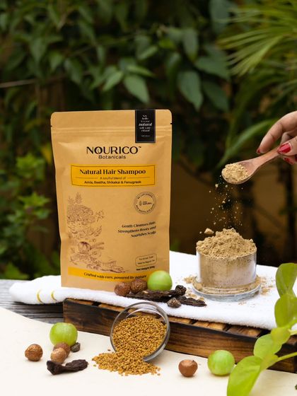 An action shot captures the fine texture of the herbal shampoo powder being sprinkled from a wooden spoon. The outdoor, natural light setting reinforces the product's connection to nature.