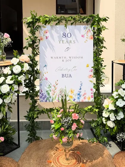 A beautiful welcome sign for an 80th birthday, celebrating "80 Years of Warmth, Wisdom & Joy." The watercolor floral design and surrounding fresh flower arrangements created a soft and welcoming entrance for guests.
