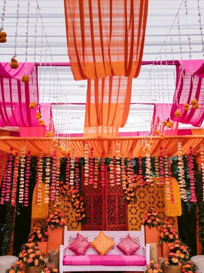 A beautiful mehendi stage decorated in shades of orange and pink. The setup features draped fabrics, hanging floral strings, and a carved backdrop for the main seating.