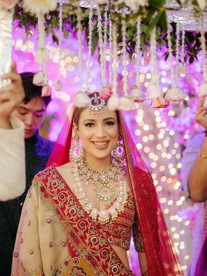 A close-up of the radiant bride during her entrance. She walks under a delicate 'phoolon ki chadar' (floral canopy), with a backdrop of soft bokeh lights creating a dreamy effect.