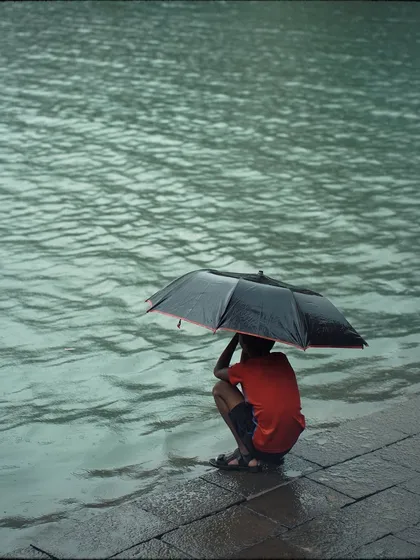 A young boy in a red shirt crouches under an umbrella at the water's edge. It's a quiet, introspective moment, capturing a child's patient observation of the rain.