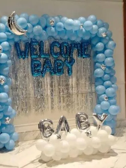 A classic "Welcome Baby" setup perfect for a home celebration. This design uses a blue balloon arch over a silver fringe curtain, with foil letters and a cloud of white balloons at the base.