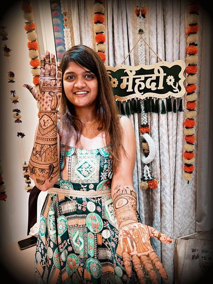 This happy bride is showing off her complete mehndi look, with detailed designs on both her hands and arms. The artwork features a mix of personal stories and traditional Indian art.