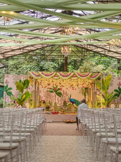 The full view of a South Indian wedding venue we designed, with a stunning peacock-themed mandap as the centerpiece. The seating is arranged to give every guest a perfect view of the ceremony, set within a beautiful glasshouse structure that blends with the surrounding greenery.