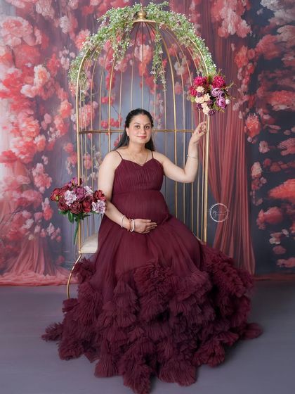 A gorgeous solo portrait in a deep maroon ruffled gown. The mom-to-be is seated in the golden birdcage, creating a dramatic and elegant scene.