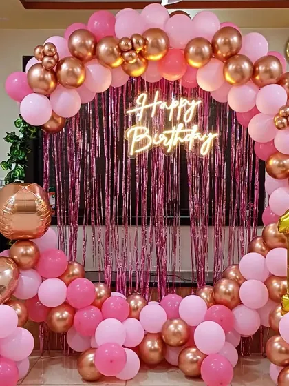 A classic first birthday decoration in pink and rose gold. The setup includes a full balloon arch over a tinsel curtain, a "Happy Birthday" neon sign, and a large number "1" foil balloon.