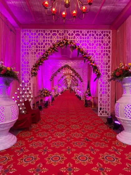 A grand and traditional entrance walkway with a red carpet, white lattice arches, floral arrangements, and rows of candle-style lights.