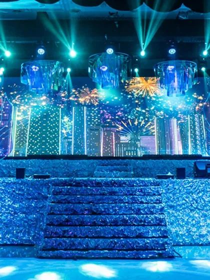 The Sangeet stage illuminated with a fireworks graphic on the LED screen. The iridescent chandeliers and textured stage front catch the light beautifully, creating a dazzling display of color and reflection.