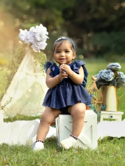 A happy smile from the birthday girl, sitting on a white crate in a park. The blue and white floral theme looks stunning against the green grass.