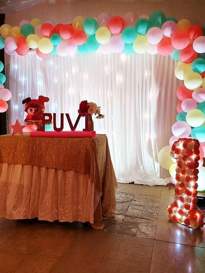 A vibrant and colorful first birthday party for Puvi. This setup includes a multi-color pastel balloon arch over a fairy-light curtain, a personalized name sign on the cake table, and a custom light-up number '1' to mark the special occasion.
