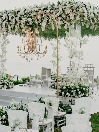 A wide view of the serene, all-white mandap setup by the sea. The combination of a lush floral canopy, elegant chandeliers, and clear acrylic guest chairs creates a look of pure, timeless sophistication.