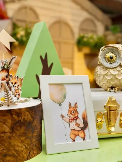 A close-up of the charming table decor at Ryne's woodland party. The details include a golden owl figurine, a framed squirrel illustration, and tiny animal cutouts on a wooden log slice.