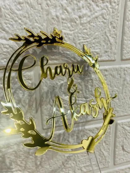 The "Charu & Akash" topper with a leafy wreath design, shown against a lighted background to highlight its golden shine.