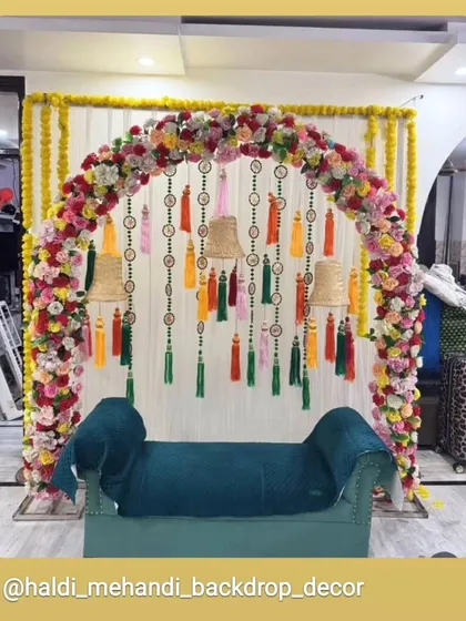 This elegant seating for a home ceremony features a beautiful floral arch with unique hanging tassels and lampshades. The teal-colored sofa adds a modern touch to the traditional decor.