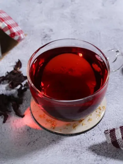 This photograph showcases a cup of hibiscus tea. The deep red color of the tea is the focal point, contrasted against a textured white background with dried hibiscus petals scattered around.