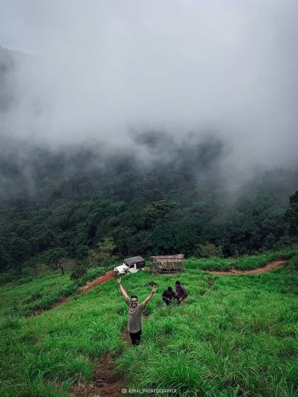 A trekker celebrating on a grassy hill in Wayanad, with a jeep and misty forests below. This is the joy of exploration.