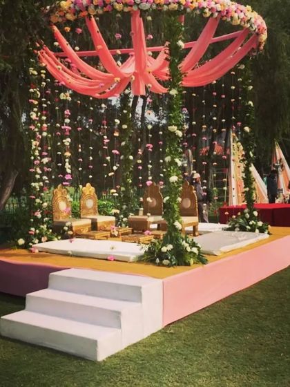 A beautiful outdoor mandap for a day ceremony. The stage is decorated with pink drapes and hanging floral strings, creating a soft and romantic ambiance.