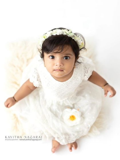 A sweet six-month-old girl in a simple white dress, looking up with wide, curious eyes. The high-key lighting creates a pure and angelic feel.