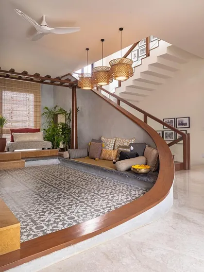 This sunken lounge area, defined by a gracefully curved wooden bench and traditional patterned tiles, creates an intimate space for conversation and connection within an open-plan layout. It’s a modern take on the traditional Indian 'baithak' or seating area.