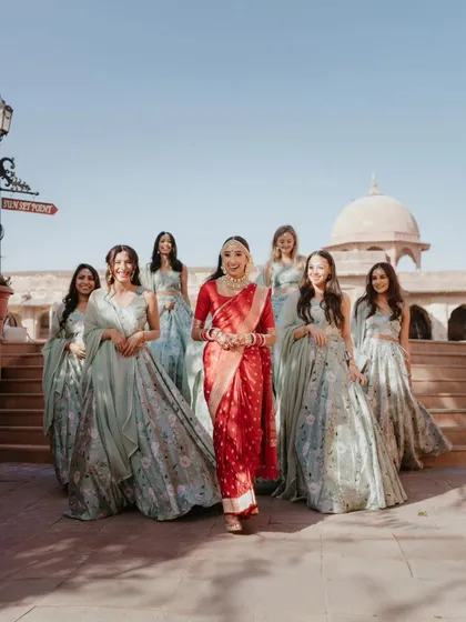 The bride, dressed in a traditional red saree for her South Indian ceremony, walking with her bridesmaids. This shot highlights the beautiful contrast of colors against the heritage property.