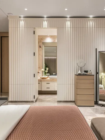 Subtle luxury defines this serene primary bedroom, featuring a hidden door to the ensuite bathroom within a fluted wall panel. The integration of natural materials like stone and warm veneer paneling creates a soothing ambiance, crafted with a blend of functionality and elegance.