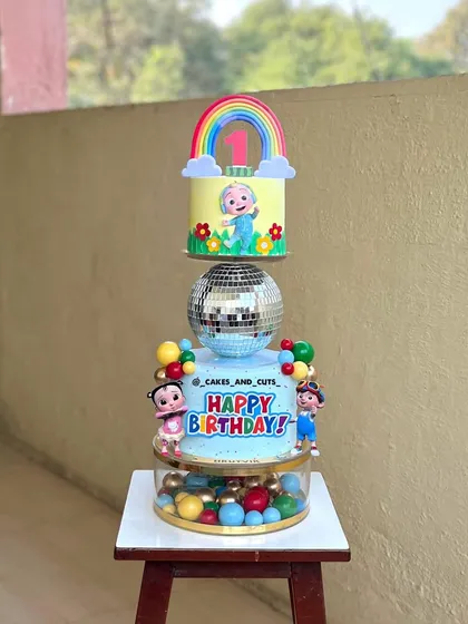 A colorful two-tier Cocomelon cake for a first birthday celebration. This design includes a disco ball separator and is decorated with characters from the popular show.