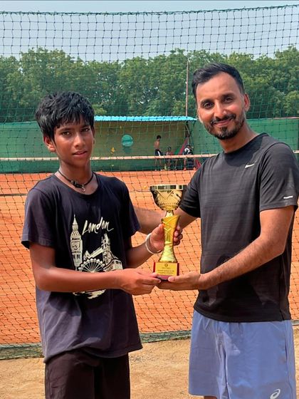 Divyansh Lekhi is awarded his trophy for winning the U18 title at a UTR tournament. He stands with his coach in front of the net on a sunny day.