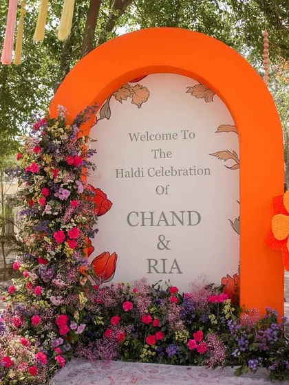 A bright and welcoming entrance for a Haldi celebration. The bold orange arch is adorned with a cascade of colorful fresh flowers, setting a cheerful tone for the event.