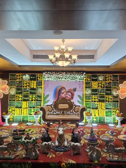 A naming ceremony stage with a Yashoda and Krishna themed backdrop, a marigold panel wall, and a central cow prop.