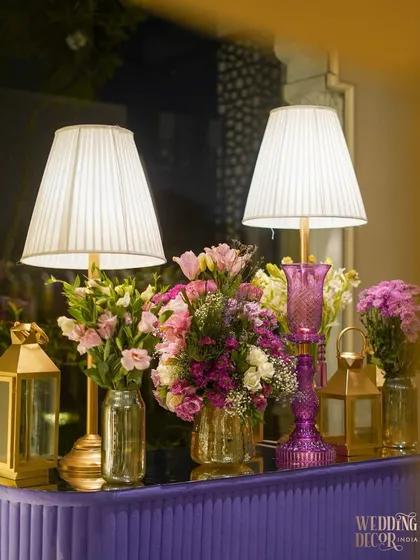 A beautiful tabletop arrangement featuring elegant lamps, golden lanterns, and bouquets of pink and purple flowers. The soft light from the lamps creates a warm and intimate dining atmosphere.