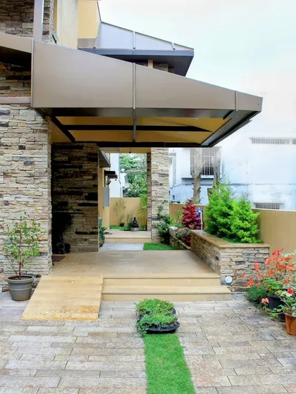 The entrance is designed to be a welcoming transition from the outside in, featuring a mix of stone pavers, a small garden bed, and a protective metal-clad overhang.