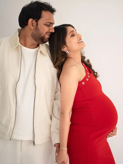 A candid and joyful portrait of an expecting couple. The mom-to-be, in a vibrant red dress, leans back against her partner with a happy smile, basking in the moment.