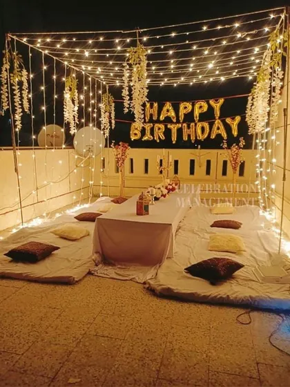 A cozy birthday dinner arrangement on a terrace, with floor seating, a canopy of fairy lights, and hanging floral decorations.