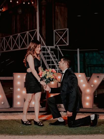 A classic kneeling proposal shot in front of the 'MARRY ME' letters, capturing the timeless romance of the moment. The focus is entirely on the couple's interaction.