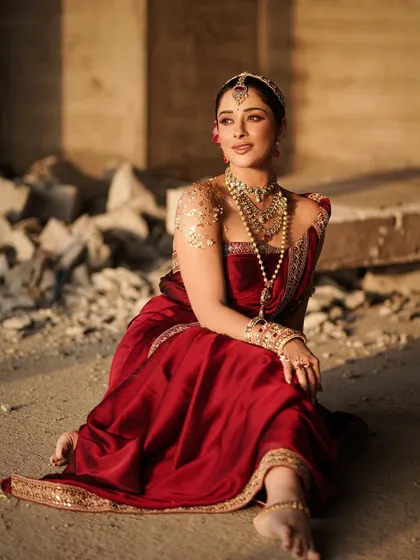 A beautiful seated pose that showcases the rich fabric of the saree and the detailed anklets. The warm, golden hour lighting enhances the richness of the red and gold tones.
