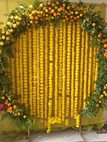 A circular backdrop for a Haldi ceremony, featuring a frame of green leaves and orange marigolds around a curtain of yellow marigold strings.