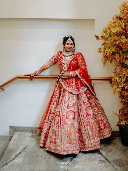 A classic full-length bridal portrait. The bride stands gracefully by a staircase, her red lehenga on full display.