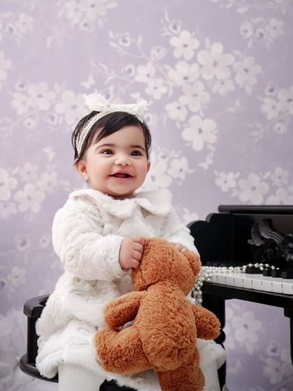 A sweet smile from this baby girl as she holds her teddy bear, sitting at a miniature piano during her milestone shoot.