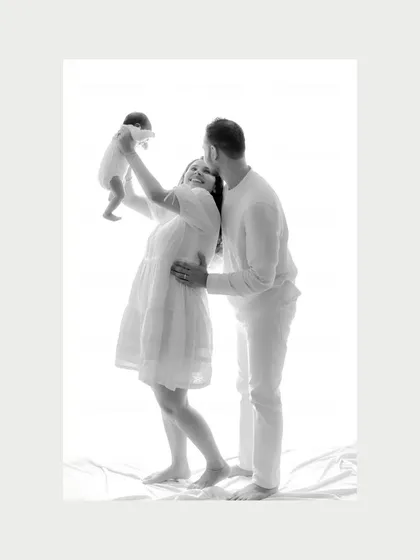 A joyful lift towards the sky. In this black and white studio shot, a mother joyfully raises her newborn as the father stands lovingly behind her. It’s a moment of celebration and happiness, beautifully captured in a timeless style.