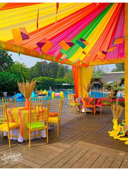 Poolside seating under a rainbow-draped canopy. The tables are set with colorful linens and simple floral centerpieces, creating a cheerful dining area by the water.