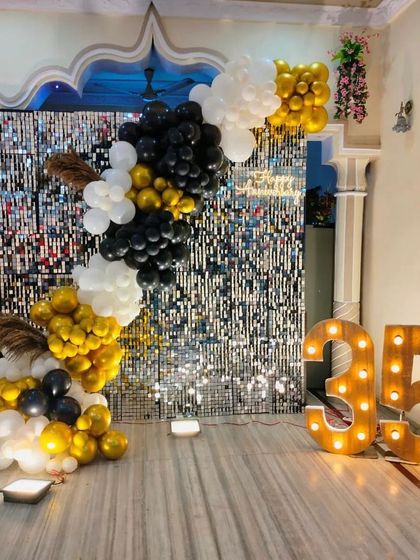 A sophisticated backdrop for a 35th birthday, featuring a silver shimmer wall that adds texture and shine. The asymmetrical balloon garland in gold, black, and white provides a modern and elegant frame, complemented by large light-up marquee numbers.