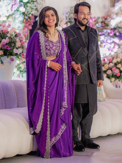 A full view of the couple's reception look. The bride's custom purple draped gown is perfectly complemented by the groom's structured black sherwani, showcasing a modern approach to wedding fashion.