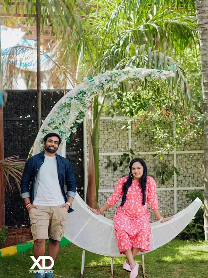 A whimsical couple's portrait featuring a large, flower-adorned crescent moon prop in a garden setting.