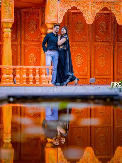 The reflection in the water perfectly mirrors the couple and the stunning orange Jaipuri-style architecture, creating a symmetrical and visually striking image.