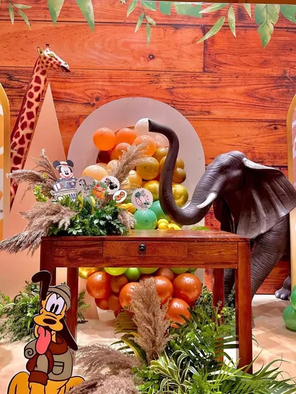 A detailed look at the jungle decor, featuring an elephant prop reaching over a wooden table. The scene is filled with character cutouts like Pluto, pampas grass, and earthy-toned balloons.