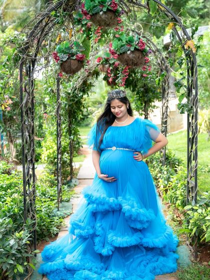 Feeling like a princess in a garden paradise. This mom-to-be wears a tiara and a stunning blue ruffled gown, standing under a floral archway.